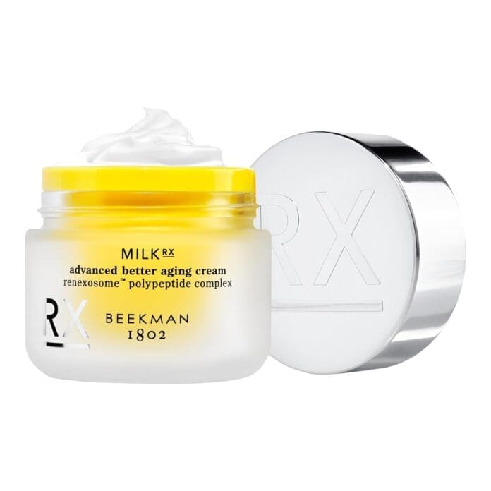 An open jar of Beekman 1802 Milk RX Advanced Better Aging Cream, revealing the creamy white texture inside. The frosted glass packaging has a yellow-tinted top, and the silver lid is embossed with the RX logo. The product's name and key technology, 