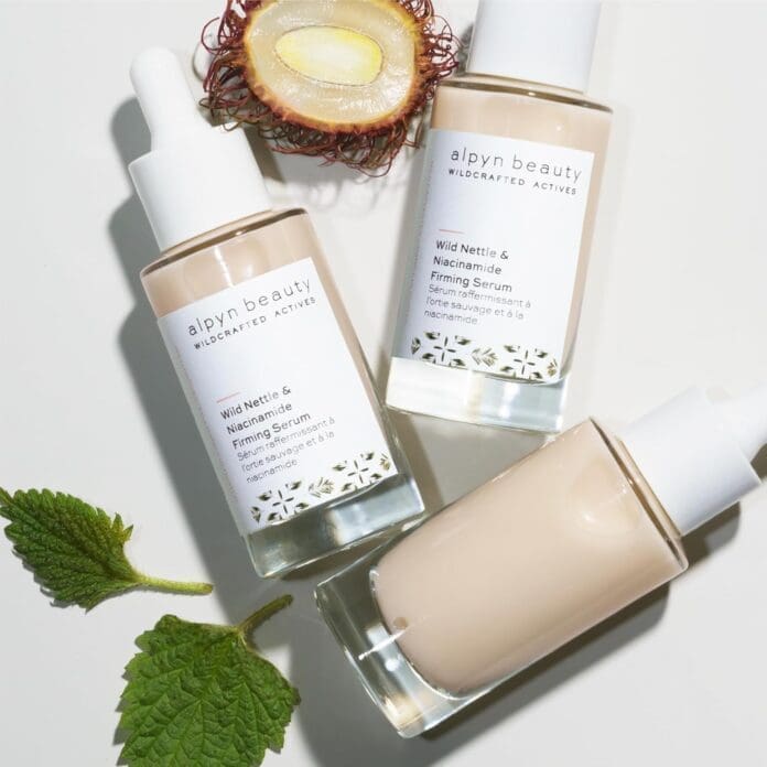 A flat lay of three bottles of Alpyn Beauty Wild Nettle & Niacinamide Firming Serum arranged on a white surface. One bottle is slightly tilted, revealing its creamy formula. The bottles have white dropper caps and minimalist labels. Surrounding the bottles are a halved rambutan fruit and fresh wild nettle leaves, indicating the product's natural ingredients.