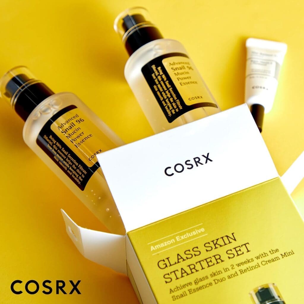 A close-up shot of the COSRX Glass Skin Starter Set on a yellow background. The set includes two bottles of Advanced Snail 96 Mucin Power Essence and a small tube of Retinol 0.1% Cream. The open box, prominently displaying the "COSRX" logo, is labeled as an "Amazon Exclusive" and highlights the promise of achieving glass skin in two weeks.

