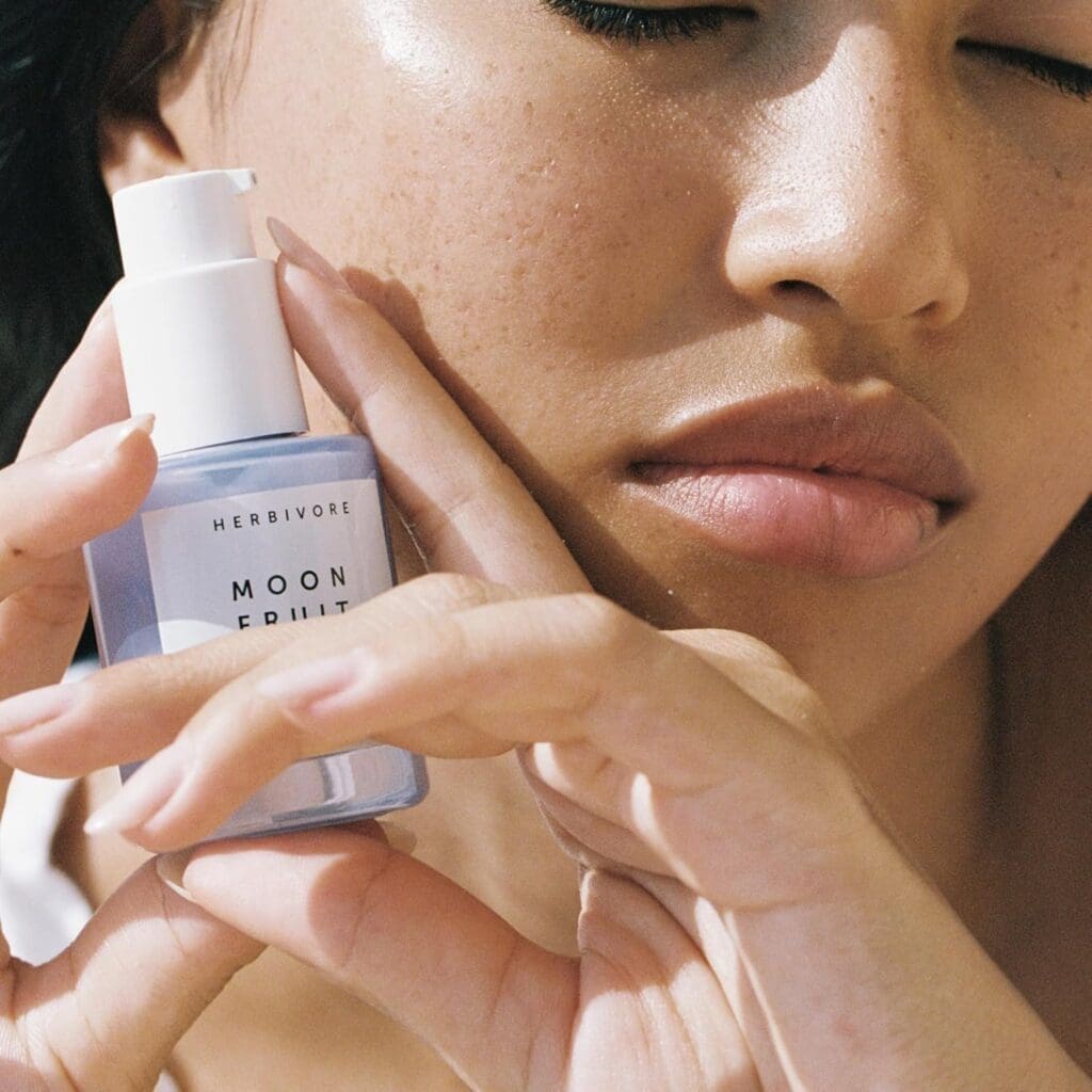 A close-up of a woman with glowing, smooth skin holding a bottle of Herbivore Botanicals' Moon Fruit Retinol Alternative Serum close to her face. She has a serene expression, and the sunlight highlights her skin’s natural texture.