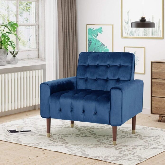 A stylish living room featuring a navy blue velvet armchair with a tufted back and seat, wooden legs with gold-tipped accents, and a modern design. The chair is placed on a light-colored patterned rug, surrounded by framed botanical artwork, a wooden dresser, and a window with sheer curtains allowing natural light.