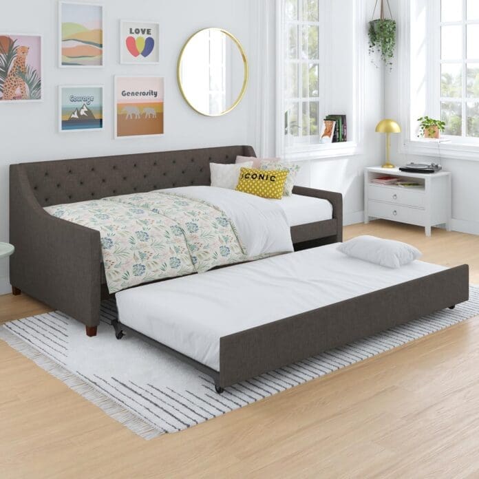 A modern upholstered daybed with a pull-out trundle bed in a stylish bedroom. The room features a contemporary design with framed wall art, a round mirror, and natural light streaming through large windows. The bed is dressed in floral bedding with decorative pillows.