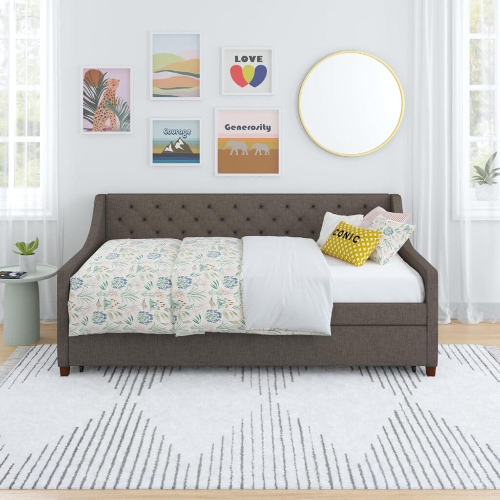 A front view of the Novogratz Her Majesty Upholstered Daybed in a well-lit bedroom. The daybed is styled with patterned bedding and colorful throw pillows, set against a backdrop of framed artwork and a round mirror. The trundle is concealed beneath the main bed for a sleek look.