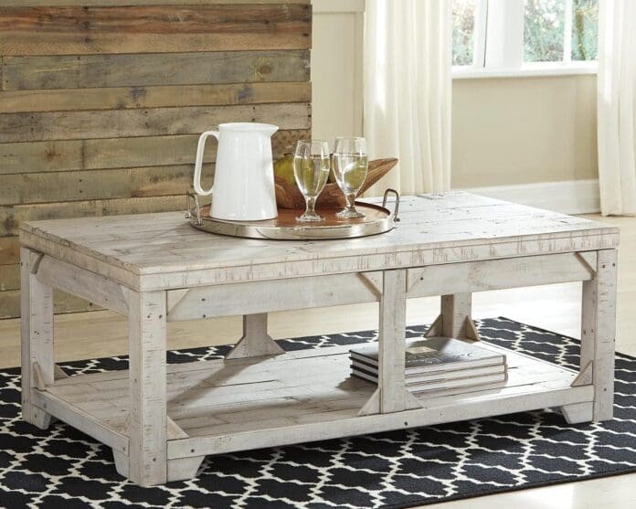 A rustic farmhouse-style lift-top coffee table with a distressed white wood finish, placed in a cozy living room with a wooden accent wall. The table features a lower shelf for storage and a lift-top surface for convenience, with a tray, glasses, and a pitcher displayed on top.