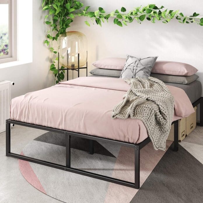 A modern bedroom featuring the ZINUS Lorelei 14-inch metal platform bed frame with a black steel structure. The bed is dressed in soft pink bedding with gray and patterned pillows, alongside a knitted blanket. Green vines hang on the white walls, and a warm-toned bedside lamp adds ambiance.